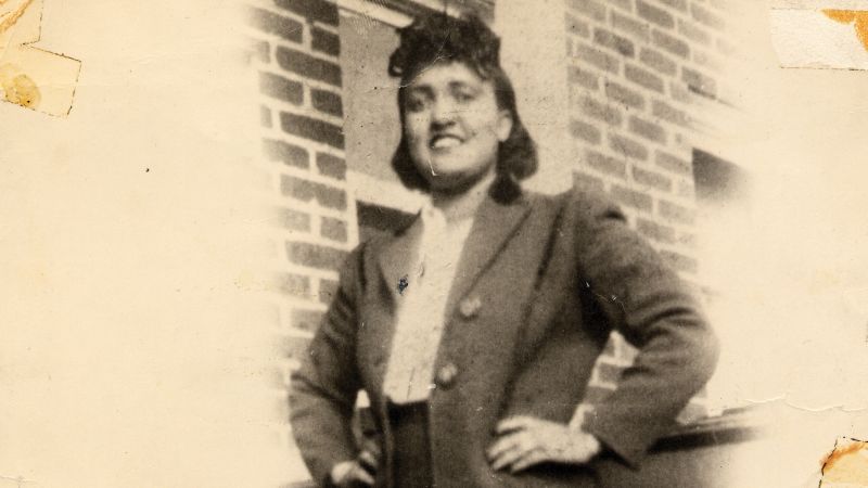 Vintage photograph of Henrietta Lacks smiling in a double-breasted coat, standing outside a brick building
